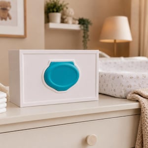 May include: A white rectangular baby wipe dispenser with a teal lid. The dispenser is on a white dresser next to a stack of diapers, a teddy bear, and a woven basket. A framed picture of a teddy bear is in the background.