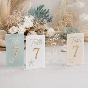 May include: Four acrylic table number signs with the word "Table" and the number "7" in gold. The signs are in various colors, including seafoam green, white, and clear, with seashell and wave designs. Perfect for a beach-themed wedding or event.