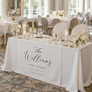May include: A white wedding table runner featuring black printed text that reads The Williams, JAMES & EMILY, and 05.24.2027 with a small heart icon. The table is decorated with white floral arrangements, tall pillar candles, and small votive candles.