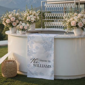 Può includere: Un bar all'aperto bianco con composizioni floreali e calici di champagne. Uno striscione con la scritta "The Cheers to Williams" è drappeggiato sul bar. Un cesto di vimini con un ombrello e fiori è sull'erba.
