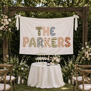 May include: A white fabric banner hanging from a wooden frame features the text THE PARKERS in large, multi-colored letters with various patterns including daisies, hearts, and checkers. Below, a round table holds a champagne tower and bottle.