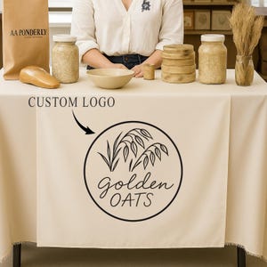 May include: A table setting featuring a beige tablecloth with a "Golden Oats" logo. The table displays a paper bag, jars, a bowl, and wooden items. The text "Custom Logo" is above the logo.