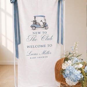 May include: A white banner with a blue golf cart illustration and text that reads "NEW TO The Club WELCOME TO Lucas Miller BABY SHOWER." The banner is held up by a white metal frame and blue ribbons. A basket of flowers sits nearby.