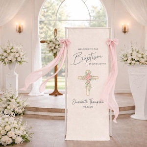 Puede incluir: Un cartel de bienvenida de bautizo color crema con el texto "Welcome to the Baptism of our Daughter" y el nombre "Elisabeth Thompson" con la fecha "06.12.26". El cartel tiene una cruz dorada con detalles florales rosas y está decorado con lazos rosas y tela fluida.