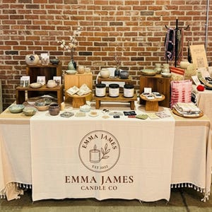 May include: A display table with various handcrafted items, including candles, pottery, and decorative objects. The table is draped with a cream-colored cloth featuring the "Emma James Candle Co" logo. The background is a rustic brick wall.