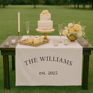 Pu&ograve; includere: Un tavolo per un ricevimento di nozze con una torta a due piani, calici di champagne e una composizione floreale. Un runner da tavolo bianco mostra "THE WILLIAMS est. 2025" in lettere nere. Sono presenti anche dessert e candele aggiuntive.