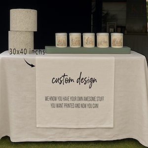 May include: A table setting featuring a white tablecloth with a custom design printed on a runner. The text on the runner reads "custom design" and "WE KNOW YOU HAVE YOUR OWN AWESOME STUFF YOU WANT PRINTED AND NOW YOU CAN!".  Two beige lampshades, each 30x40 inches, are stacked. Five small jars with illustrations are on a green shelf.
