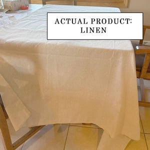 Pu&ograve; includere: Una tovaglia di lino bianca drappeggiata su un tavolo di legno. Il testo "ACTUAL PRODUCT: LINEN" &egrave; visualizzato in un rettangolo bianco. Sedie di legno circondano il tavolo, con una finestra visibile sullo sfondo. La tovaglia &egrave; di lino.