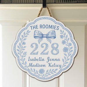 Peut inclure: Panneau de porte blanc et bleu avec un motif floral, le texte "THE ROOMIES" et le numéro "228". Le panneau comprend également les noms "Isabella Jenna Madison Kelsey". Le panneau est suspendu à un crochet noir avec une corde.