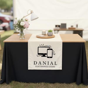 May include: A table with a black skirt and a light-colored wooden top. A cream-colored table runner displays a black graphic of a computer and smartphone, with the text "DANIAL ELECTRONICS STORE". A bouquet of flowers and a desk lamp are on the table.