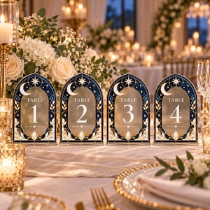 May include: Four arched table number signs featuring a dark blue celestial design with a white crescent moon, stars, and gold leaf accents. Each sign displays the word TABLE followed by the numbers 1, 2, 3, and 4 in a white serif font.