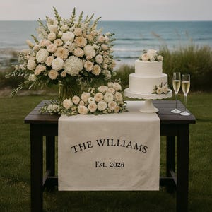 May include: A two-tiered white wedding cake with floral accents sits on a white cake stand. Champagne flutes and floral arrangements are on a dark wood table with a linen runner that reads "THE WILLIAMS Est. 2026".