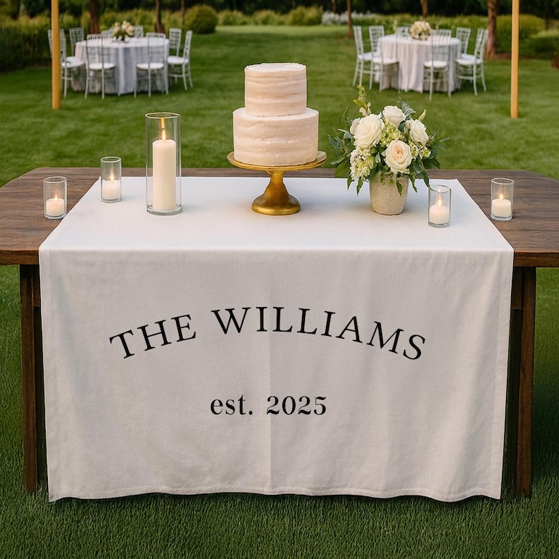 Pu&ograve; includere: Un tavolo da ricevimento nuziale con un runner bianco con la scritta "THE WILLIAMS est. 2025". Una torta a due piani su un supporto dorato, candele e composizioni floreali.