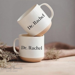 May include: Two white ceramic mugs with a tan base and handle are stacked. The text "Dr. Rachel" is printed in black on the front of each mug. The mugs are on a wooden surface.