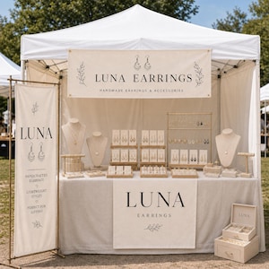 May include: A white pop-up canopy tent booth for Luna Earrings featuring a large banner with the text LUNA EARRINGS, a vertical banner with LUNA, and a table display with multiple jewelry stands, earring cards, and a wooden jewelry storage box.