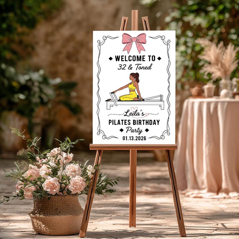May include: A white sign on a wooden easel with the text "WELCOME TO 32 & Toned" and "Leila's PILATES BIRTHDAY Party 01.13.2026". The sign features a pink bow and an illustration of a person doing Pilates. A basket of pink flowers is in the foreground.