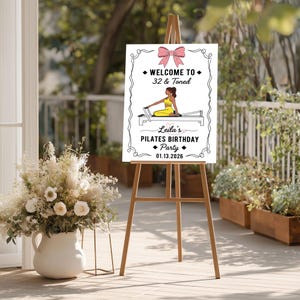 May include: A white sign on a wooden easel with the text "WELCOME TO 32 & Toned" and "Leila's PILATES BIRTHDAY Party 01.13.2026". The sign features a pink bow and an illustration of a person doing Pilates.