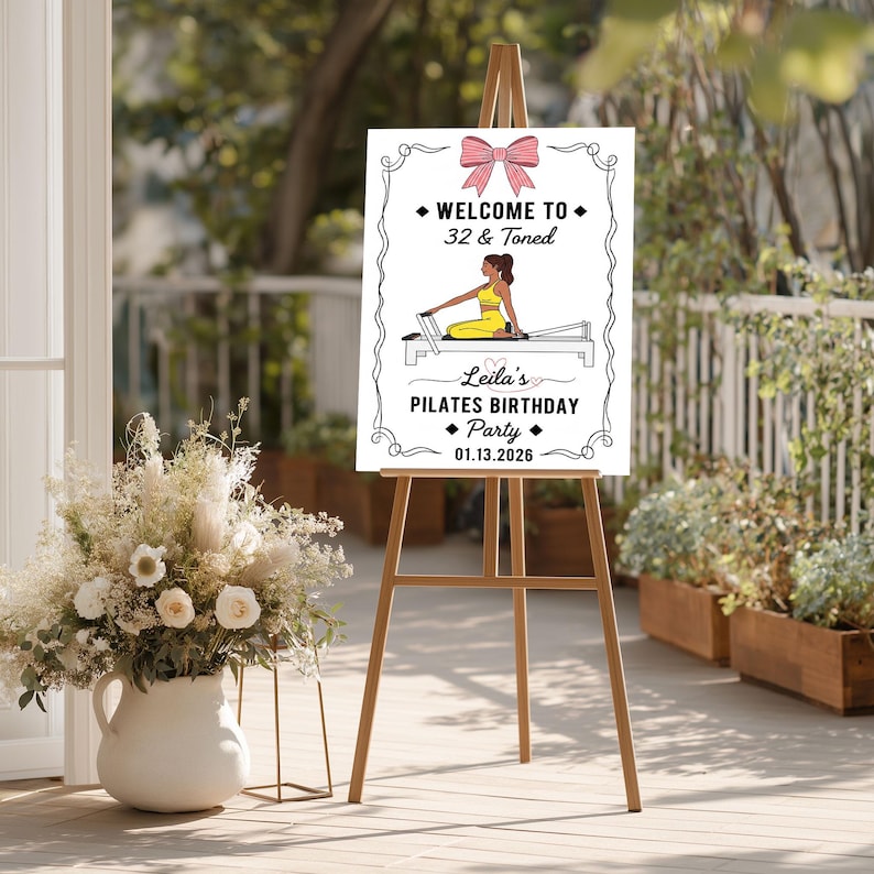 May include: A white sign on a wooden easel with the text "WELCOME TO 32 & Toned" and "Leila's PILATES BIRTHDAY Party 01.13.2026". The sign features a pink bow and an illustration of a person doing Pilates.