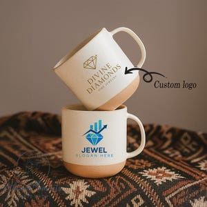 May include: Two white ceramic mugs with a tan base and handle. One mug displays the text "DIVINE DIAMONDS FINE JEWELRY" with a diamond graphic in gold. The other mug has a blue diamond graphic with the text "JEWEL SLOGAN HERE".