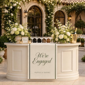 Puede incluir: Una barra de eventos de color crema con un cartel colgante que presenta líneas verticales de color verde oscuro y el texto "We're Engaged" seguido de "OLIVIA & DANIEL". La barra está decorada con dos grandes arreglos florales blancos.