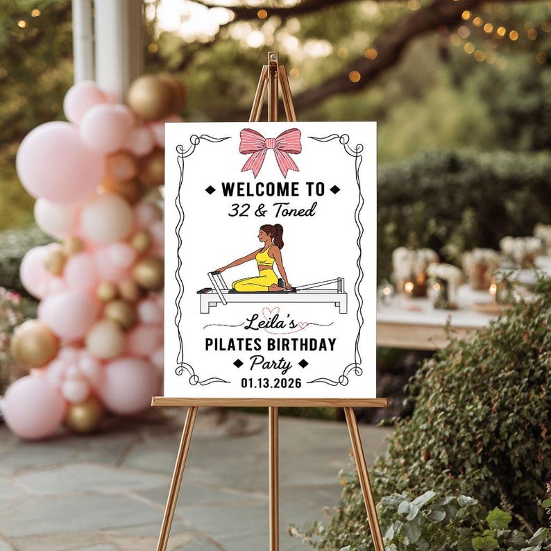May include: A white sign on a wooden easel with the text "Welcome to 32 & Toned" and "Leila's Pilates Birthday Party 01.13.2026". The sign features a pink bow and an illustration of a person doing Pilates.