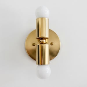 May include: A brass wall sconce featuring two white globe light bulbs. The sconce has a circular backplate and cylindrical gold-coloured fixtures. The light bulbs are positioned above and below the central fixture.