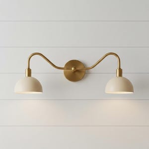 Könnte beinhalten: Wandleuchte mit zwei cremefarbenen, kuppelförmigen Schirmen. Die Leuchte hat eine Messingausführung, mit einem gebogenen Arm, der die Schirme mit einer kreisförmigen Rückplatte verbindet. Zur Wandmontage konzipiert.