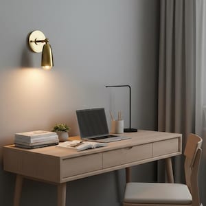 Może przedstawiać: Złota lampa ścienna z kloszem w kształcie stożka oświetla drewniane biurko. Na biurku znajduje się laptop, książki i mała roślina. Biurko ma szuflady i jasne wykończenie. Obecna jest również czarna lampa biurkowa.