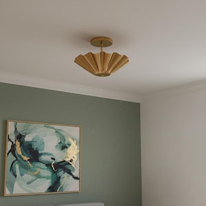Puede incluir: Una lámpara de techo dorada de estilo art déco. La luz tiene un diseño festoneado en forma de abanico y está montada en un techo blanco. La lámpara es el punto focal de la habitación, con una pintura en la pared.