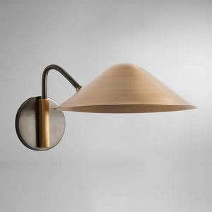 Op de afbeelding: Een wandlamp met een conische, messingkleurige kap. De lamp heeft een donkere, gebogen arm en een cirkelvormige basis met een messing accent. Het ontwerp is modern en minimalistisch, geschikt voor binnenverlichting.