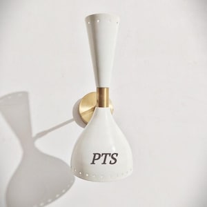 Può includere: Applique da parete bianca con un accento in ottone e un paralume conico. Il paralume riporta le lettere "PTS". L'applique è montata su una parete bianca e ha un'ombra.