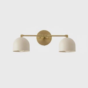 May include: A two-light vanity light fixture with a gold finish. The fixture has two off-white, dome-shaped shades and a horizontal bar connecting them. The light is mounted on a round gold backplate.