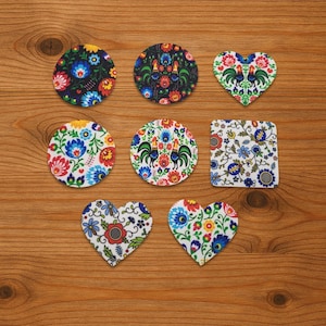 Può includere: Una collezione di sottobicchieri colorati e fantasia in varie forme, tra cui cerchi, cuori e un quadrato. I disegni presentano motivi floreali e di galli in vivaci tonalità di blu, rosso, giallo e verde, su sfondo bianco o scuro.