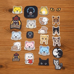 May include: A selection of cat-themed stickers in diverse designs and colours. Features stickers with cat faces, cat eyes, and cats in various poses. Some stickers include text, such as "SPY CAT" and "STAY FRESH".