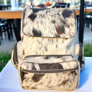 May include: A backpack with a cowhide pattern in black, brown, and white. The bag has a flap closure, two zippered pockets, and adjustable straps. The backpack is made of a soft, textured material.