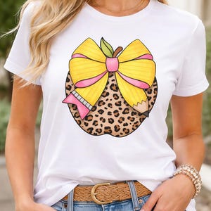 May include: White t-shirt featuring a graphic of an apple with a yellow bow, pink ribbon, and leopard print. A pink pencil and green leaf are also part of the design. The shirt is a casual, everyday wear item.
