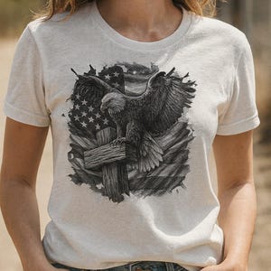 Puede incluir: Camiseta gris claro con un diseño gráfico en blanco y negro de un águila sobre una cruz de madera, con la bandera estadounidense de fondo. La camiseta tiene mangas cortas y cuello redondo.