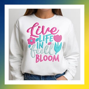 Puede incluir: Sudadera blanca con la frase bordada "Live Life in Full Bloom" en rosa, turquesa y blanco. El diseño incluye una flor rosa, un tulipán rosa y hojas turquesas. La sudadera es de cuello redondo.