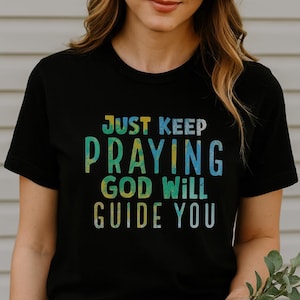 May include: Black t-shirt featuring the message "JUST KEEP PRAYING GOD WILL GUIDE YOU" in a colourful, distressed font. The tee has a classic crew neck.