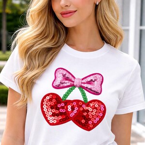 May include: White t-shirt with a sequined cherry design. The cherries are red and heart-shaped, with a pink sequined bow and green stem. The shirt has a classic crew neck.