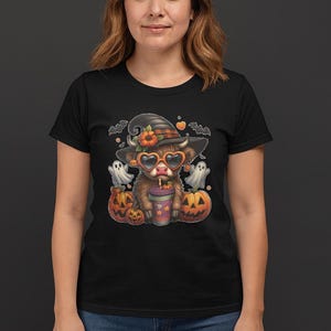 May include: Black t-shirt featuring a cartoon Highland cow wearing a witch hat, heart-shaped sunglasses, and holding a drink. The cow is surrounded by pumpkins, ghosts, and bats, creating a Halloween theme.