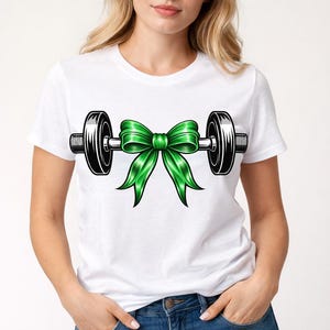 May include: White t-shirt with a barbell design, featuring a bright green bow in the middle. The barbell has black weights and a silver bar. The shirt is worn by a person in blue jeans.