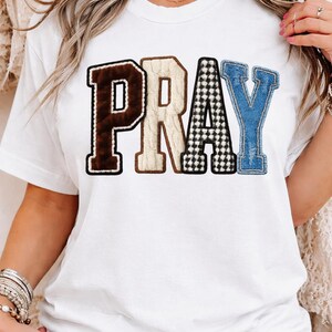 Peut inclure: T-shirt blanc avec le mot "PRAY" dans un motif patchwork. La lettre "P" est marron avec une texture floue, "R" est beige, "A" est à carreaux noirs et blancs, et "Y" est en denim bleu.
