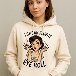 May include: Cream-colored hoodie with the text "I Speak Fluent Eye Roll" and a cartoon illustration of a woman rolling her eyes. The hoodie has a drawstring hood and a front pocket.
