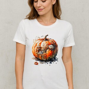 May include: White t-shirt featuring a watercolor illustration of a pumpkin with a spooky house and ghost inside. The pumpkin is orange, brown, and green, with a dark background. A small pumpkin is at the bottom. The shirt is a crew neck.