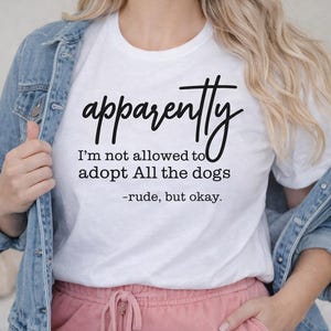 Può includere: T-shirt bianca con la scritta "apparently I'm not allowed to adopt All the dogs - rude, but okay." La maglietta è abbinata a una giacca di jeans blu e pantaloni rosa. Il testo sulla maglietta è in un carattere scritto a mano.