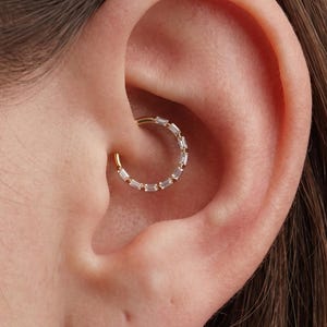 Peut inclure: Une boucle d'oreille créole circulaire en or ornée d'une série de petites pierres transparentes rectangulaires taillées en baguette, disposées le long du bord extérieur. L'anneau est placé dans le cartilage interne de l'oreille.