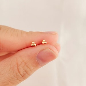 Könnte beinhalten: Ein Paar kleine, goldfarbene Ohrstecker, jeweils mit drei winzigen, kugelförmigen Perlen, die in einem dreieckigen Cluster angeordnet sind. Die Ohrringe werden zwischen Fingern vor einem weichen, cremefarbenen Hintergrund gehalten.