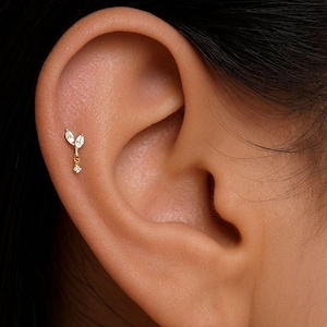 Puede incluir: Primer plano de una oreja con un pendiente dorado. El pendiente presenta dos piedras claras con corte marquesa y un pequeño diamante colgante. Una delicada pieza de joyería.