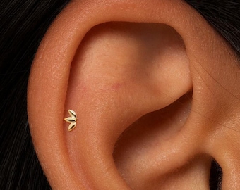 May include: A small, gold-coloured earring in the shape of a three-petal flower. The earring is shown close-up, set against the backdrop of a person's ear and dark hair.